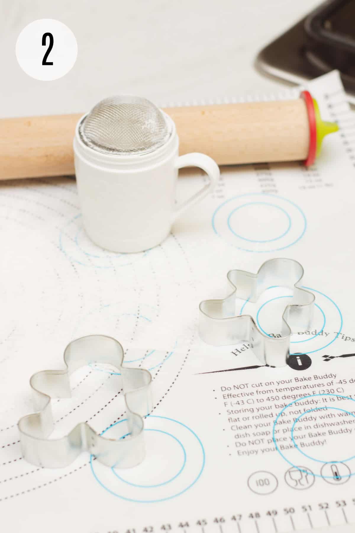 Printed baking mat with gingerbread man cookie cutters, powdered sugar/flour shaker and wooden rolling pin. 