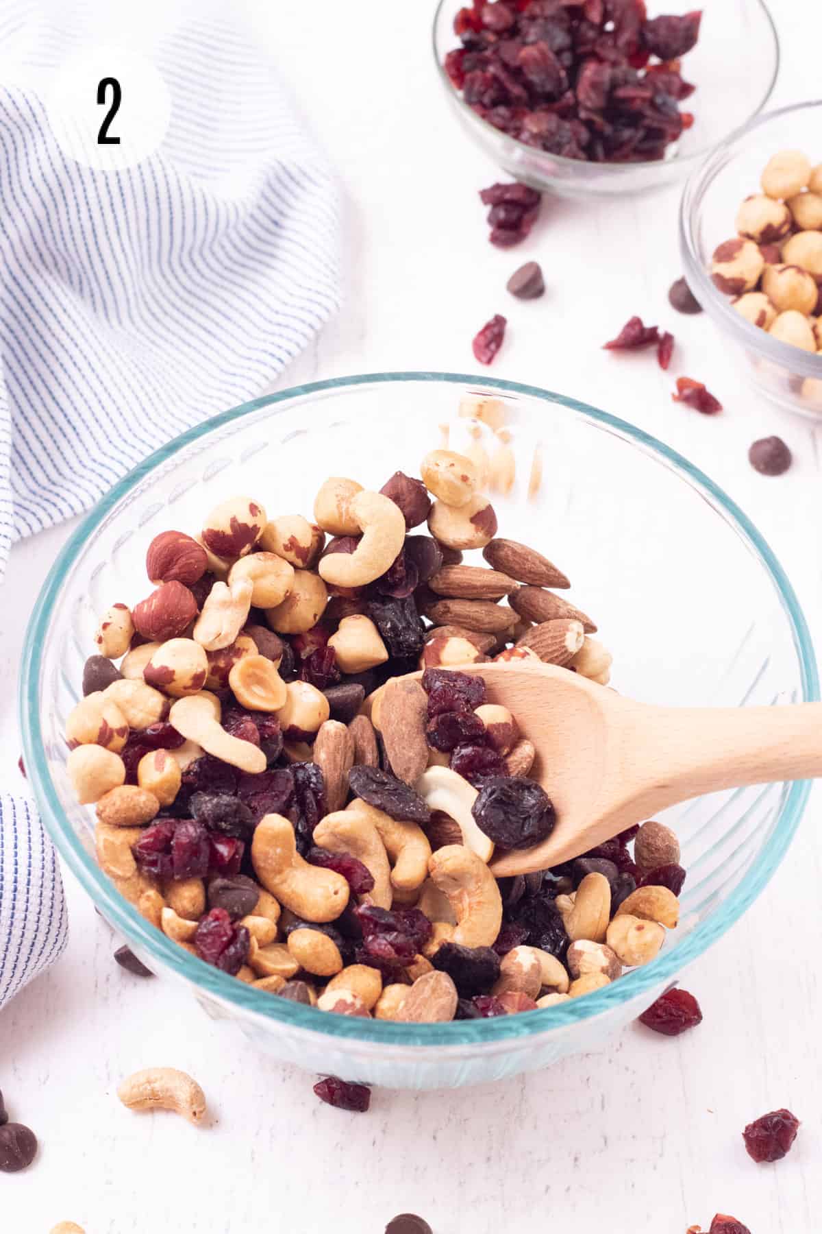 Glass bowl with wooden spoon stirring mixture of dried fruit, roasted nuts and chocolate chips and smaller bowls of ingredients in background with blue and white striped towel. 