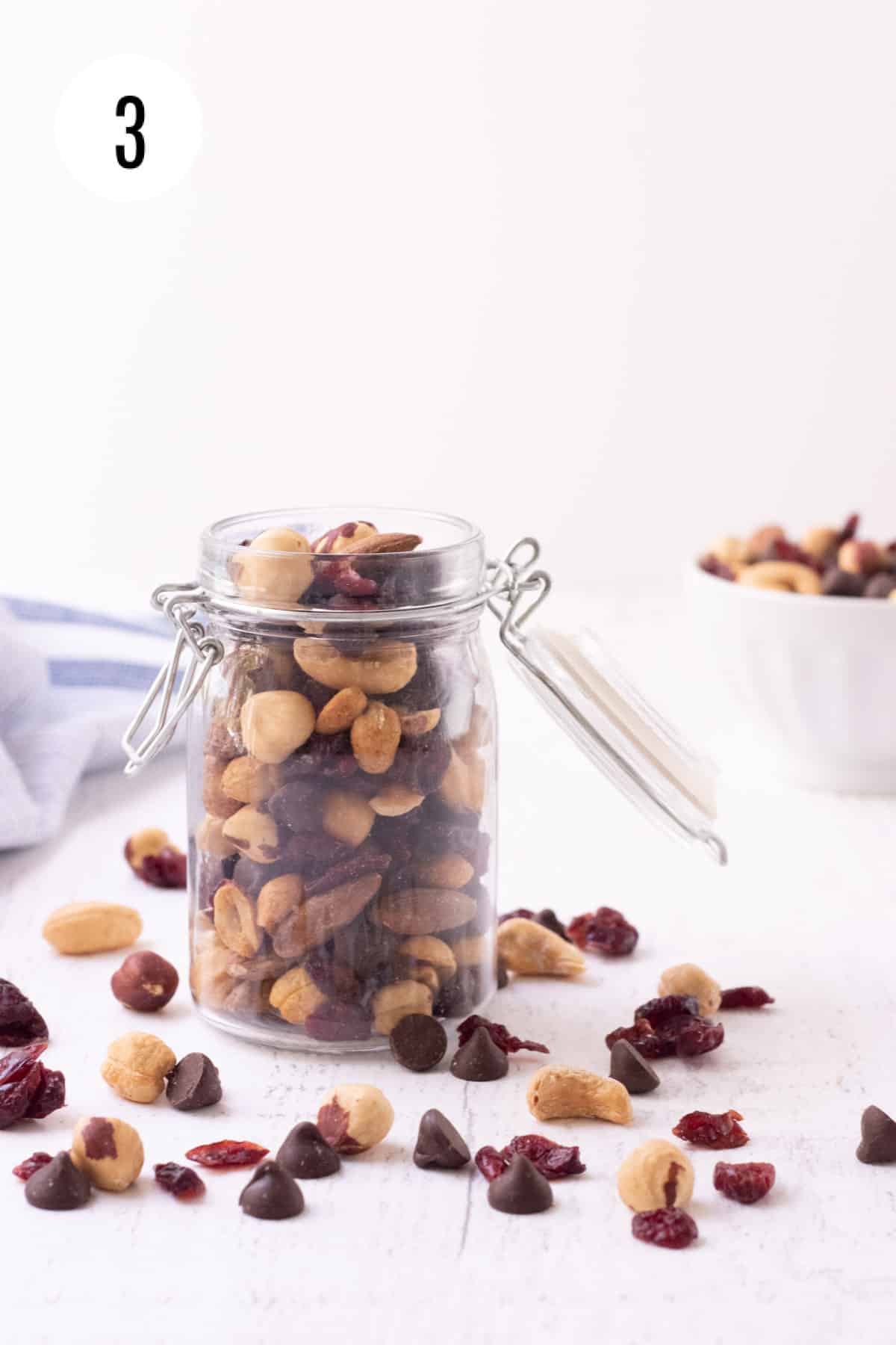 Glass jar with dark chocolate cherry trail mix and ingredients in foreground with white bowl of trail mix in background. 