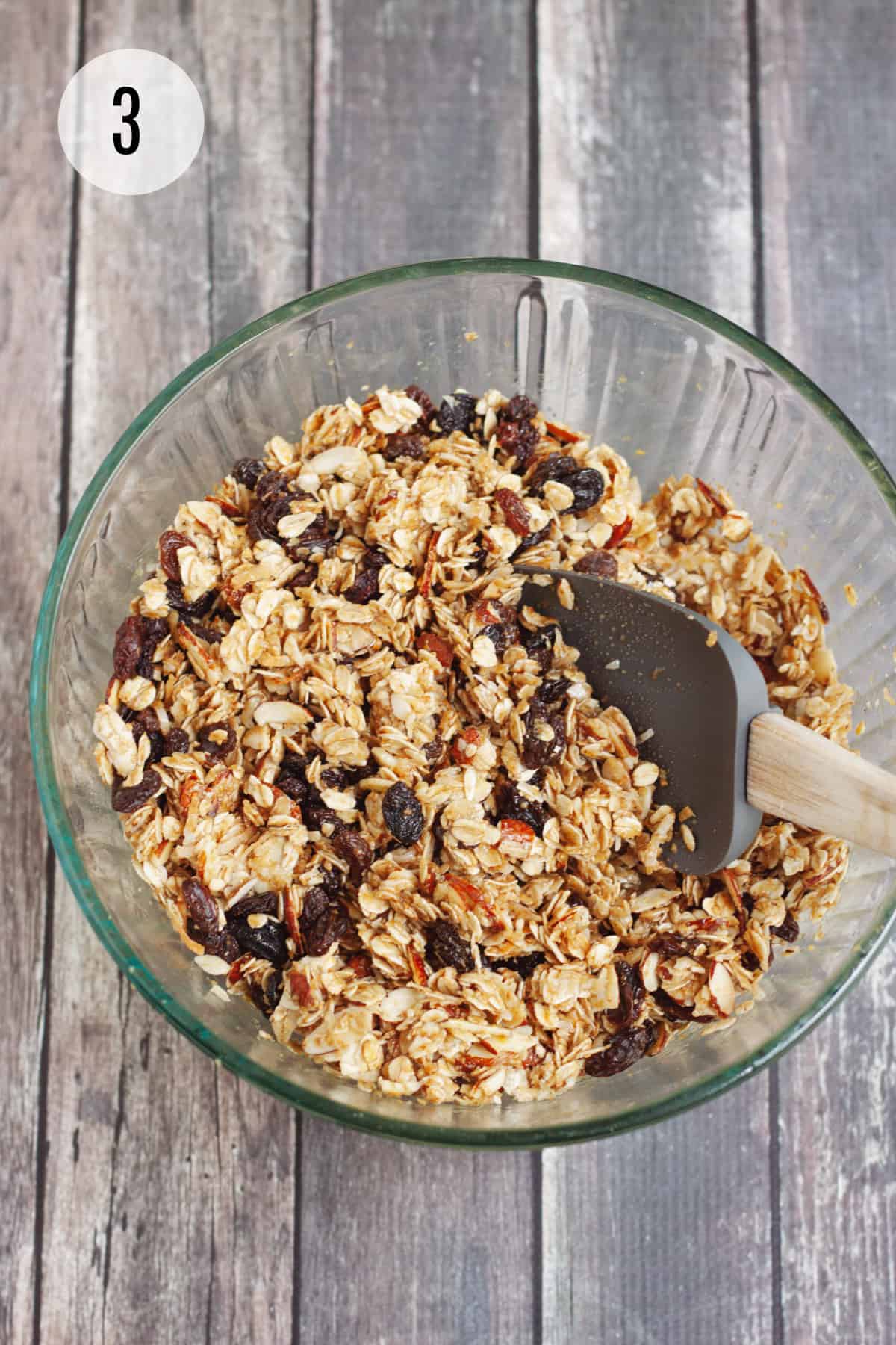 Glass bowl with homemade granola bar ingredients being stirred by grey rubber spatula. 