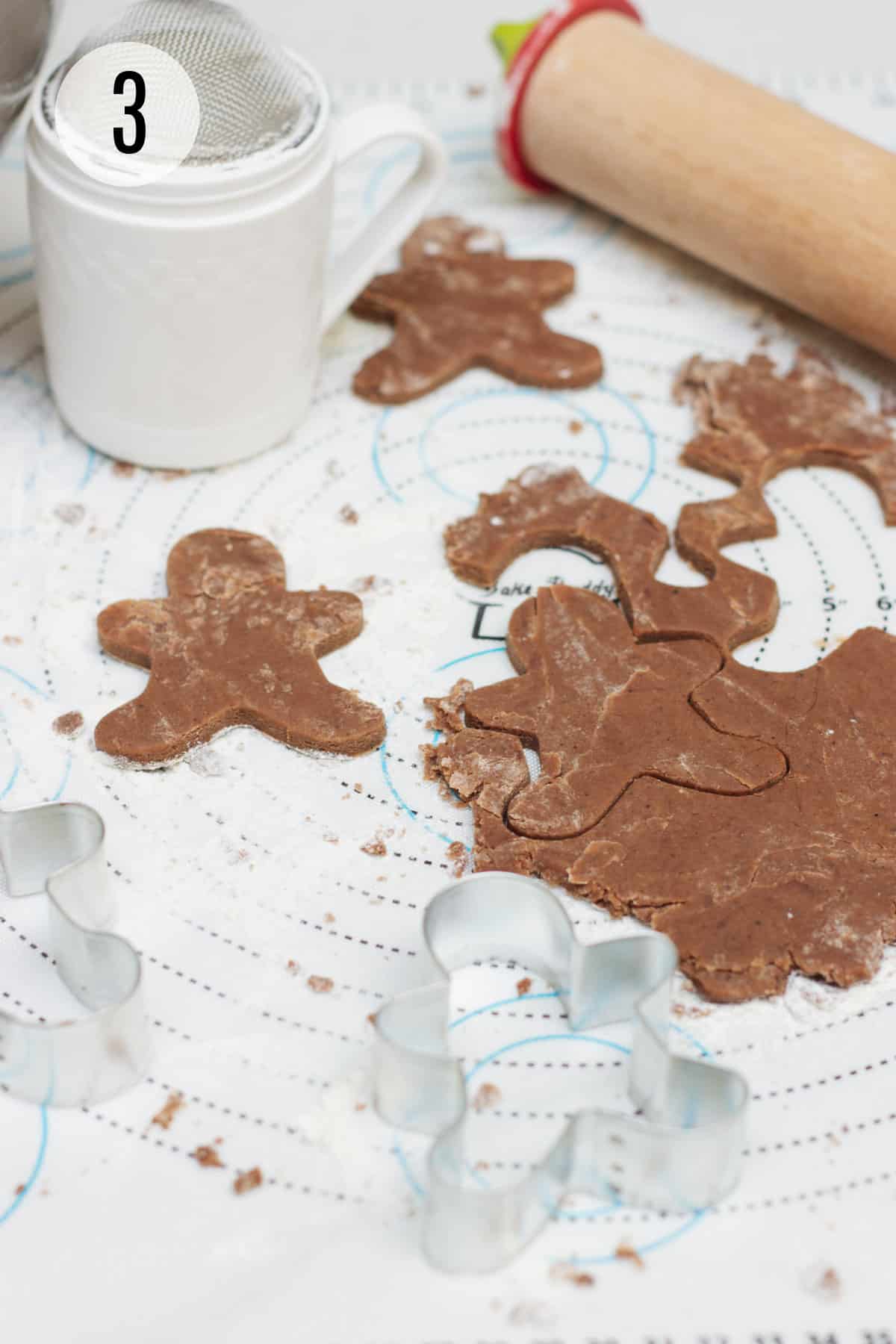 Mexican chocolate cut out sugar cookie dough on a baking mat cut into gingerbread man shape with flour/powdered sugar sifter and wooden rolling pin in upper background and metal cookie cutter in foreground. 