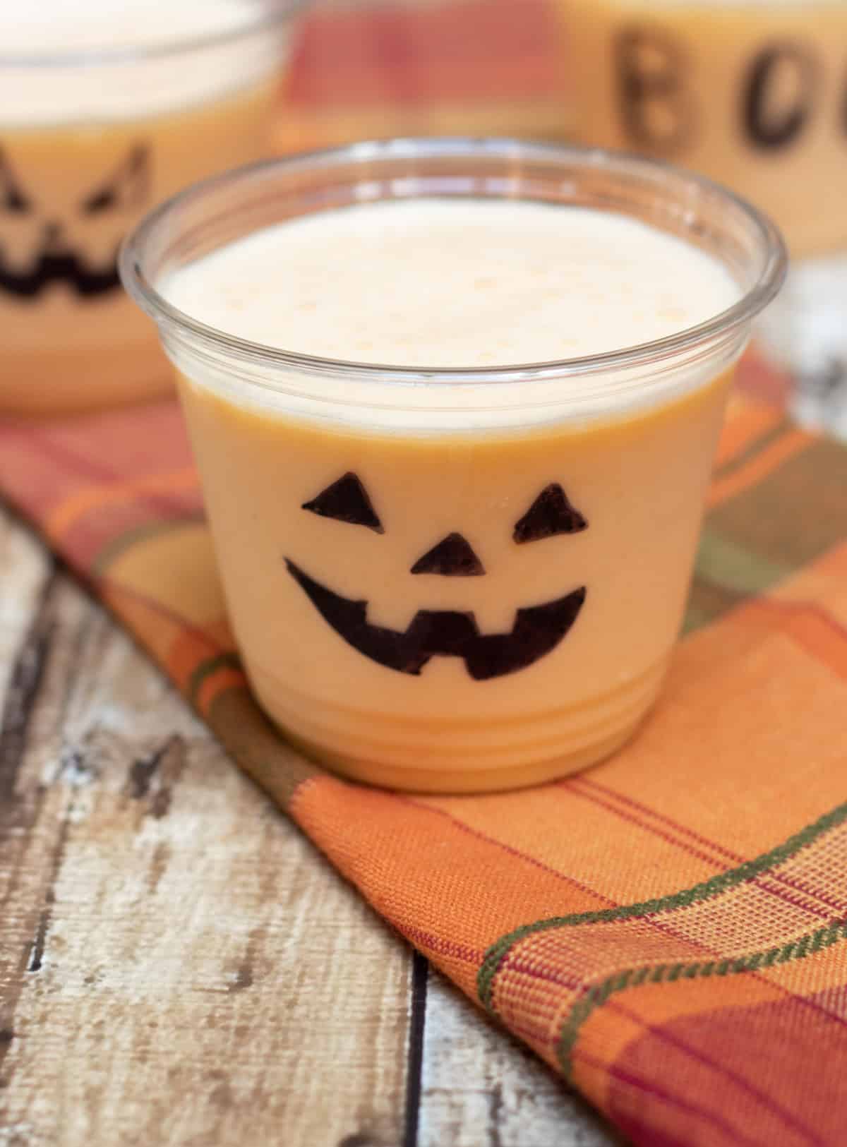 Clear cup decorated with jack-o-lantern face with orange peach smoothie on an orange plaid napkin. 