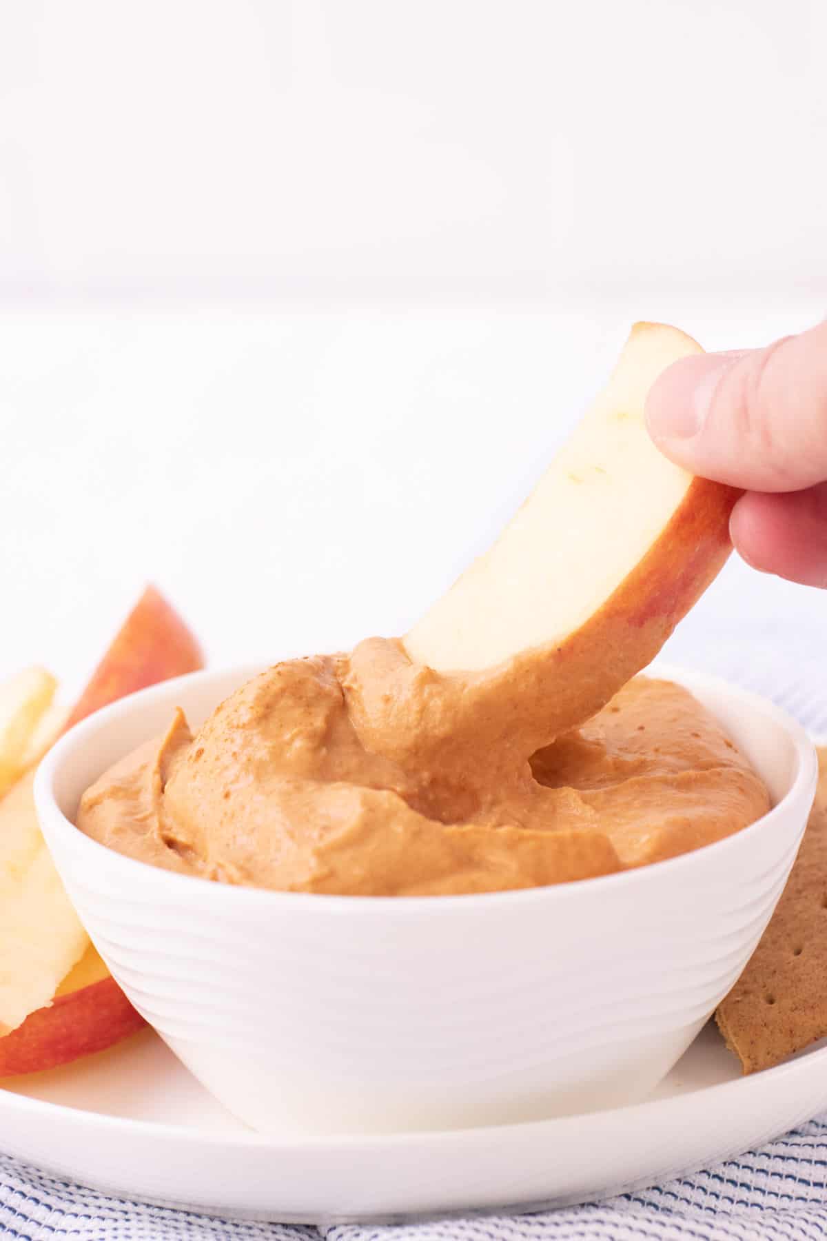 White bowl of pumpkin cheesecake dip with hand dipping an apple slice into dip with apple slices and graham crackers on plate in background on a blue and white striped towel. 