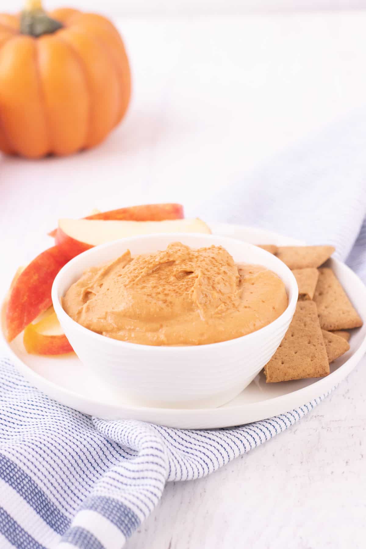 White bowl with creamy pumpkin cheesecake dip sprinkled with cinnamon on a plate with apple slices and graham crackers and pumpkin in upper left corner.