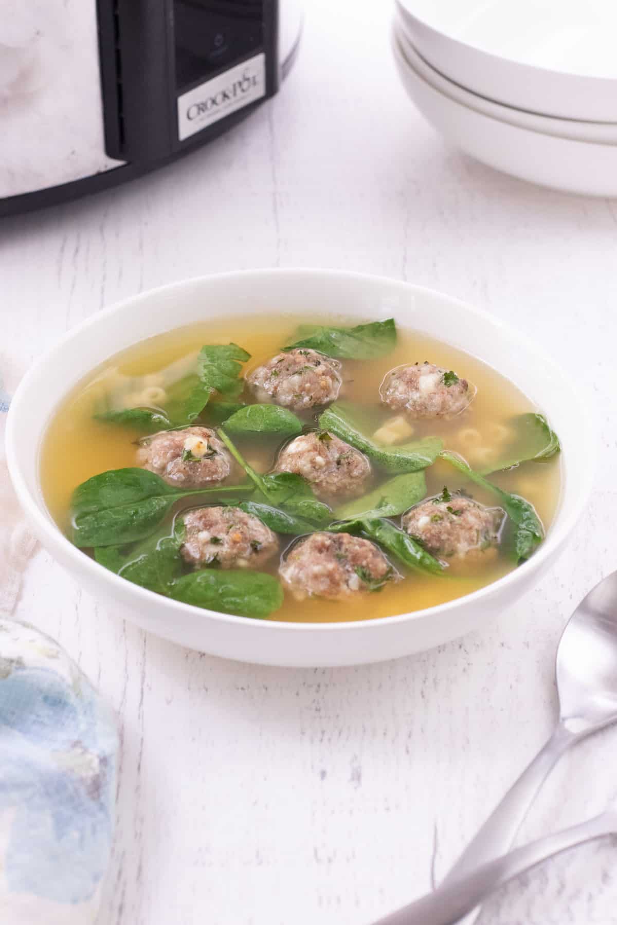 White bowl of soup containing chicken broth, spinach leaves, mini meatballs and small pasta with stack of white bowls and a slow cooker in the upper background. 