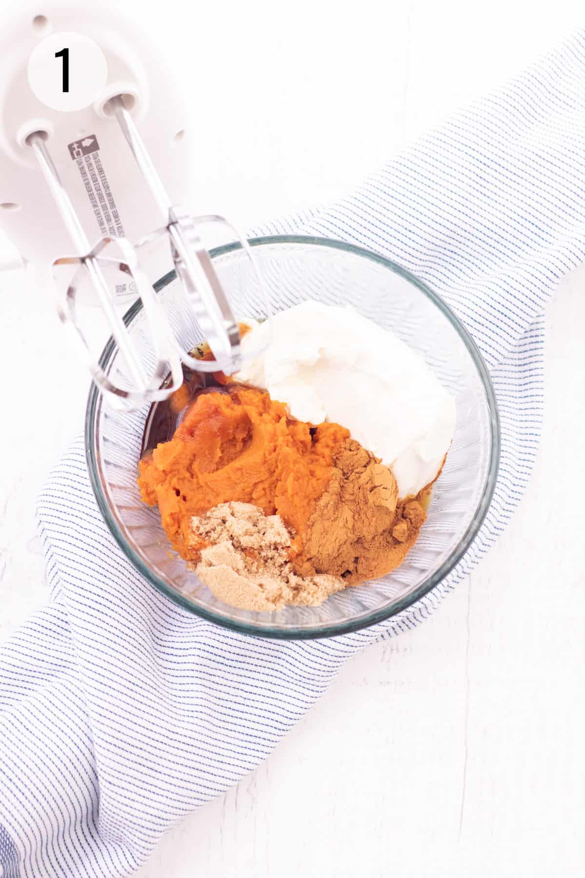 Pureed pumpkin, cream cheese, brown sugar, maple syrup and cinnamon in a glass bowl on a blue and white striped towel with electric hand mixer in upper left corner. 