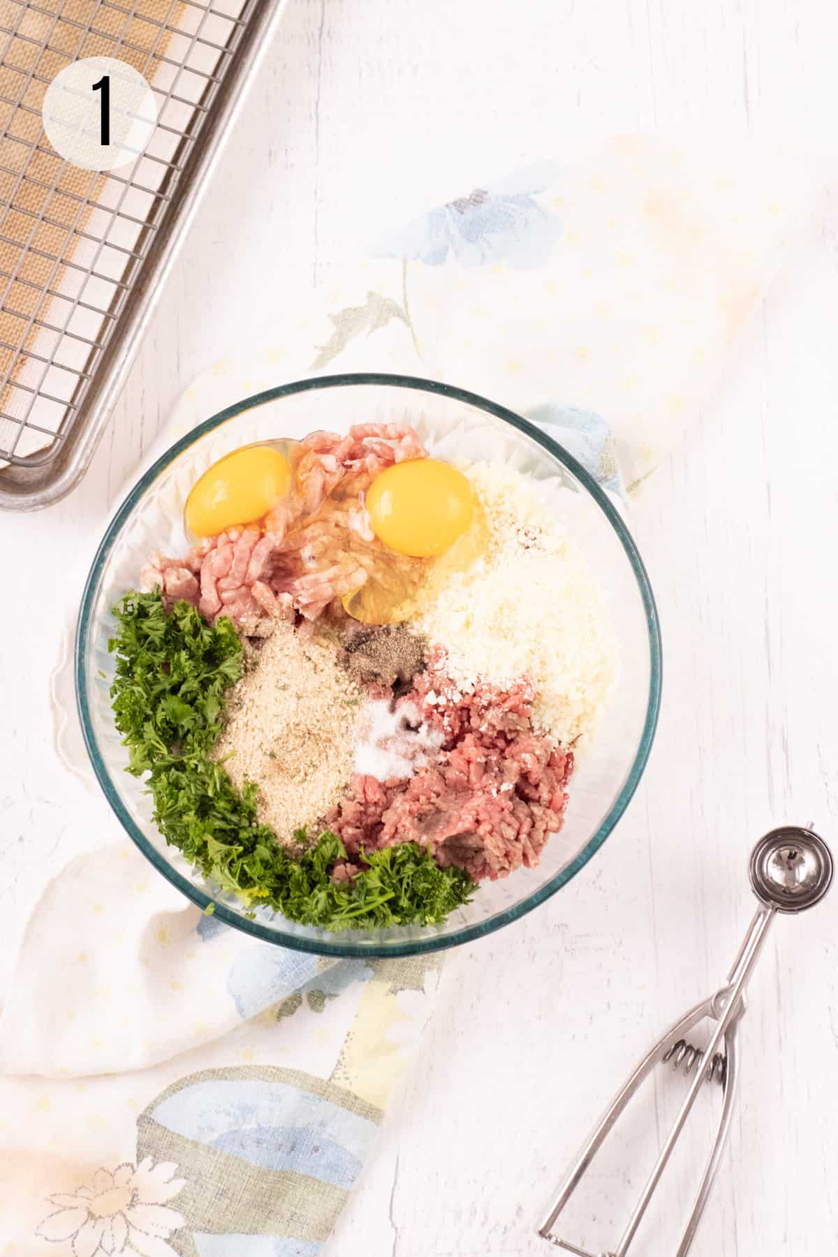 Glass bowl with ingredients for making meatballs with a small silver scoop to lower right and baking rack and pan to upper left on a light blue flowered towel. 