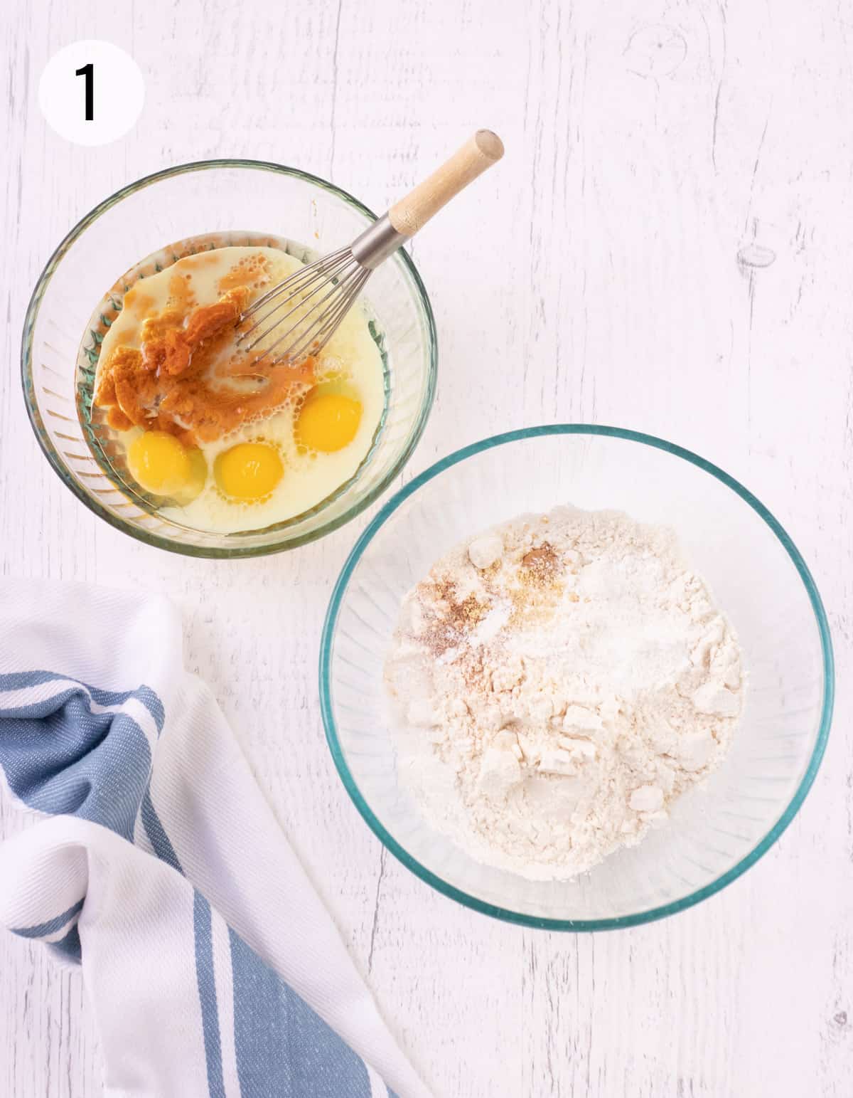 Clear glass bowls with pumpkin puree, eggs and milk in smaller bowl and flour and spice mixture in larger bowl with blue and white striped towel in lower corner. 