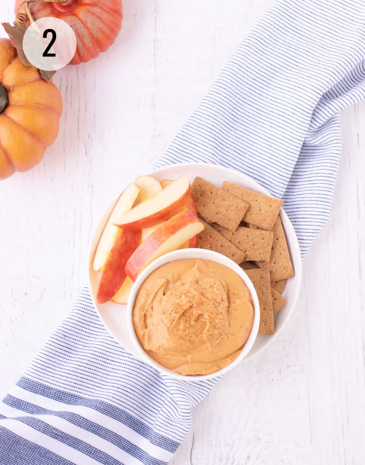 Top view of a bowl of pumpkin cheesecake dip sprinkled with ground cinnamon on a plate with apple slices and graham crackers on a blue and white striped towel with pumpkins in upper left corner. 