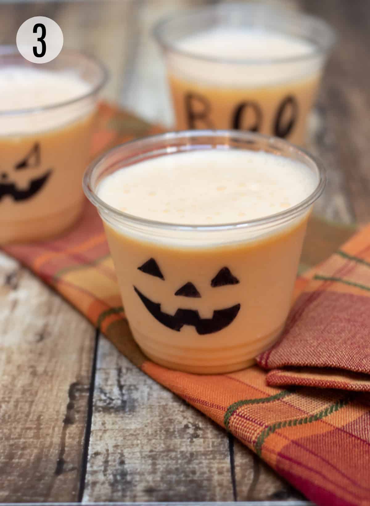 Clear cups decorated with jack-o-lantern faces and Halloween sayings filled with orange peach smoothie on an orange plaid napkin. 