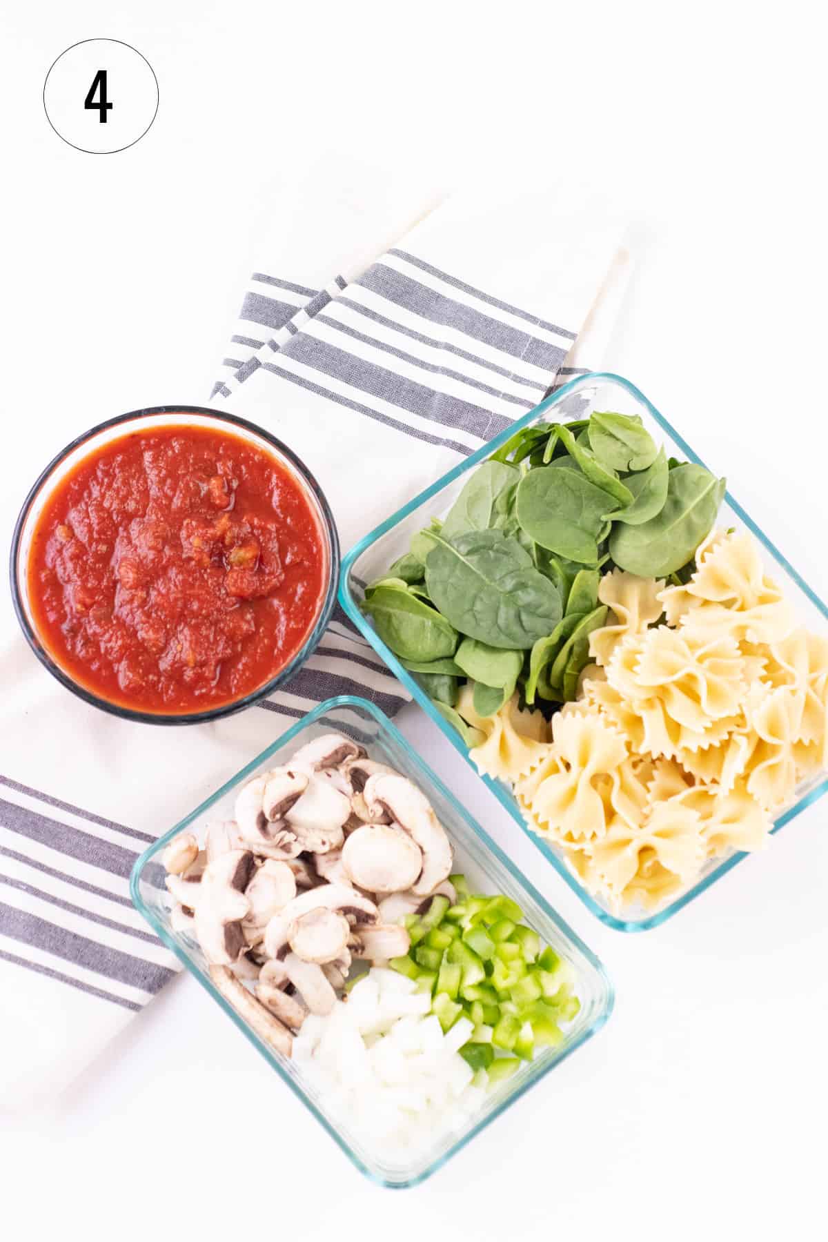 Glass containers filled with (clockwise from left) marinara sauce, baby spinach, cooked bow-tie pasta, diced green bell peppers and onions and sliced mushrooms on a striped towel to make stovetop lasagna.