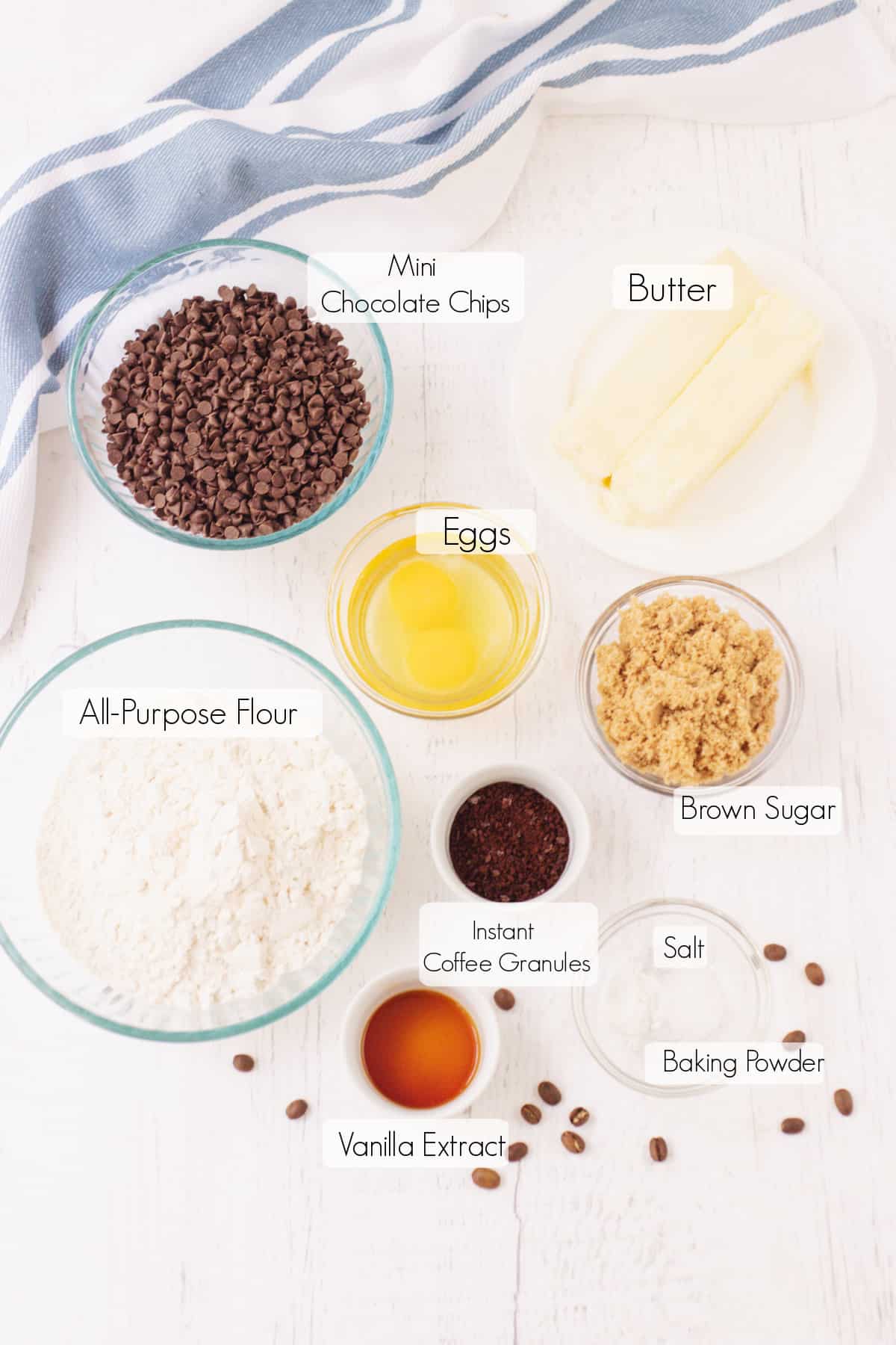 Labeled ingredients to make chocolate chip coffee cookies with blue and white towel in upper background.