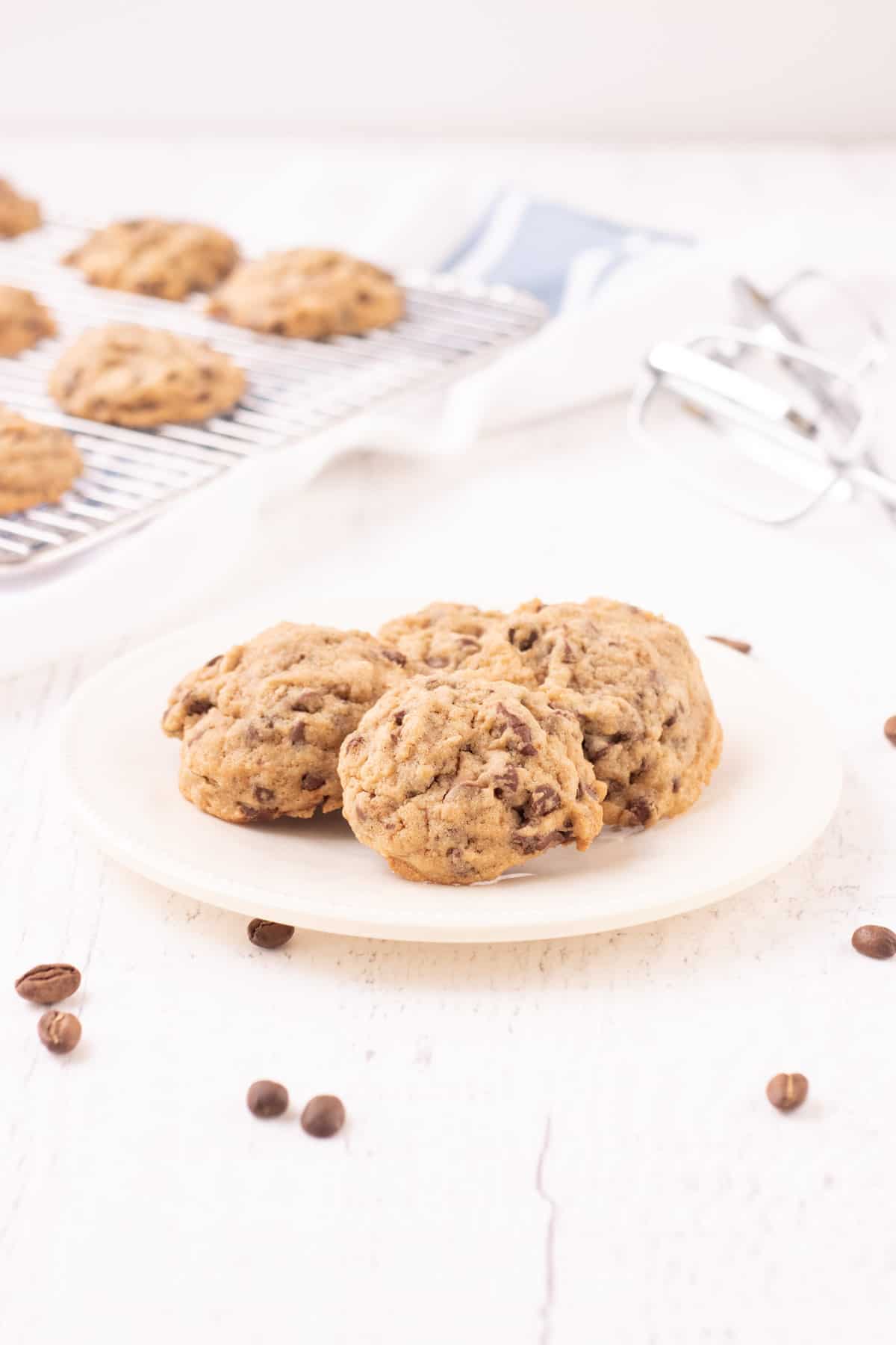 Plate of small chocolate chip coffee cookies with coffee beans sprinkled in foreground and cooling rack with cookies and mixer beater in upper background. 