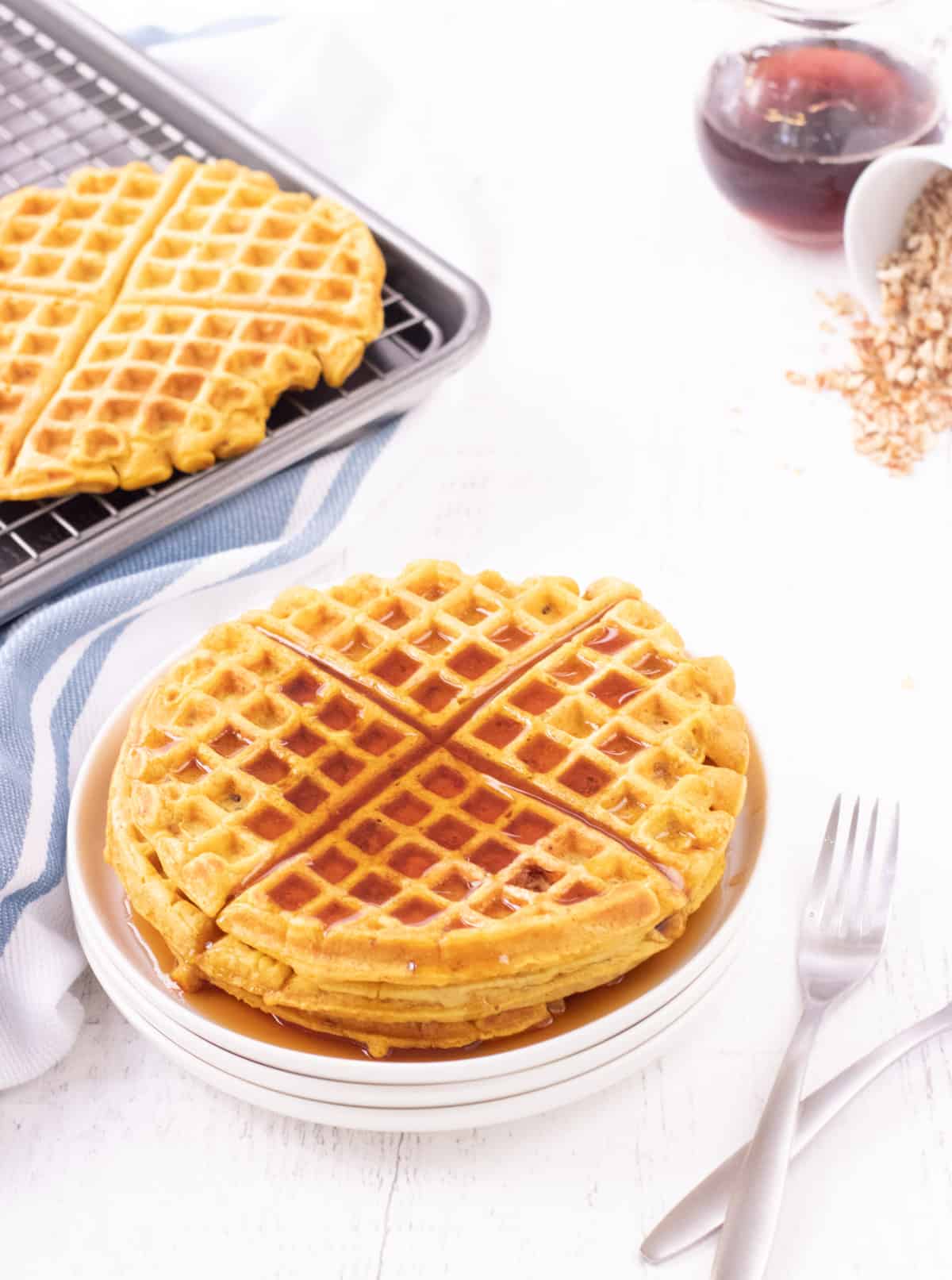 White plate of stacked pumpkin waffles with maple syrup on top and forks to side with baking sheet and rack of extra waffles and syrup and chopped nuts in upper backgrounds. 