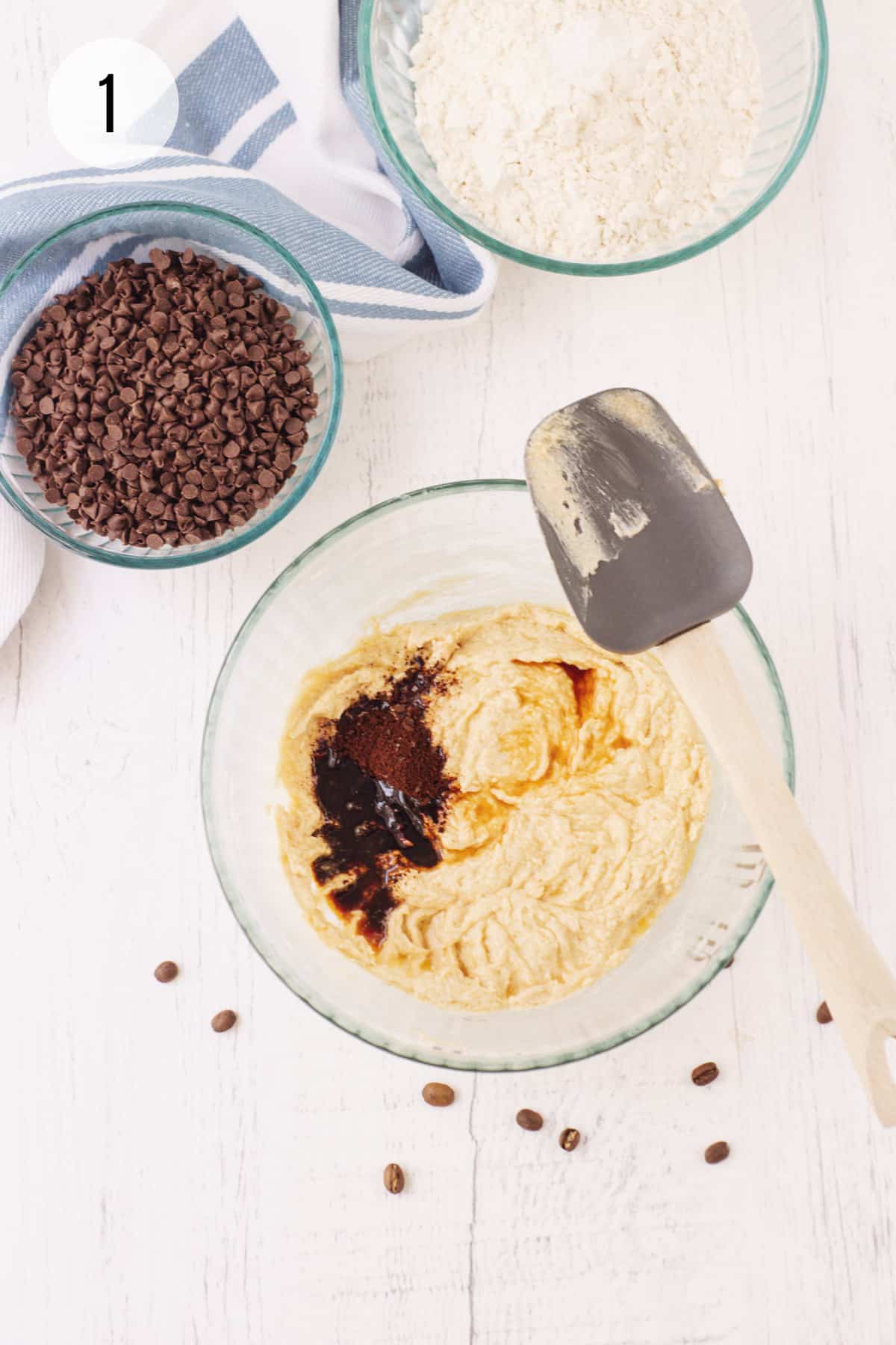 Clear bowl with creamed butter and sugar with vanilla extract and instant coffee granules and grey and wooden rubber spatula with bowls of chocolate chips and flour mixture in upper background.