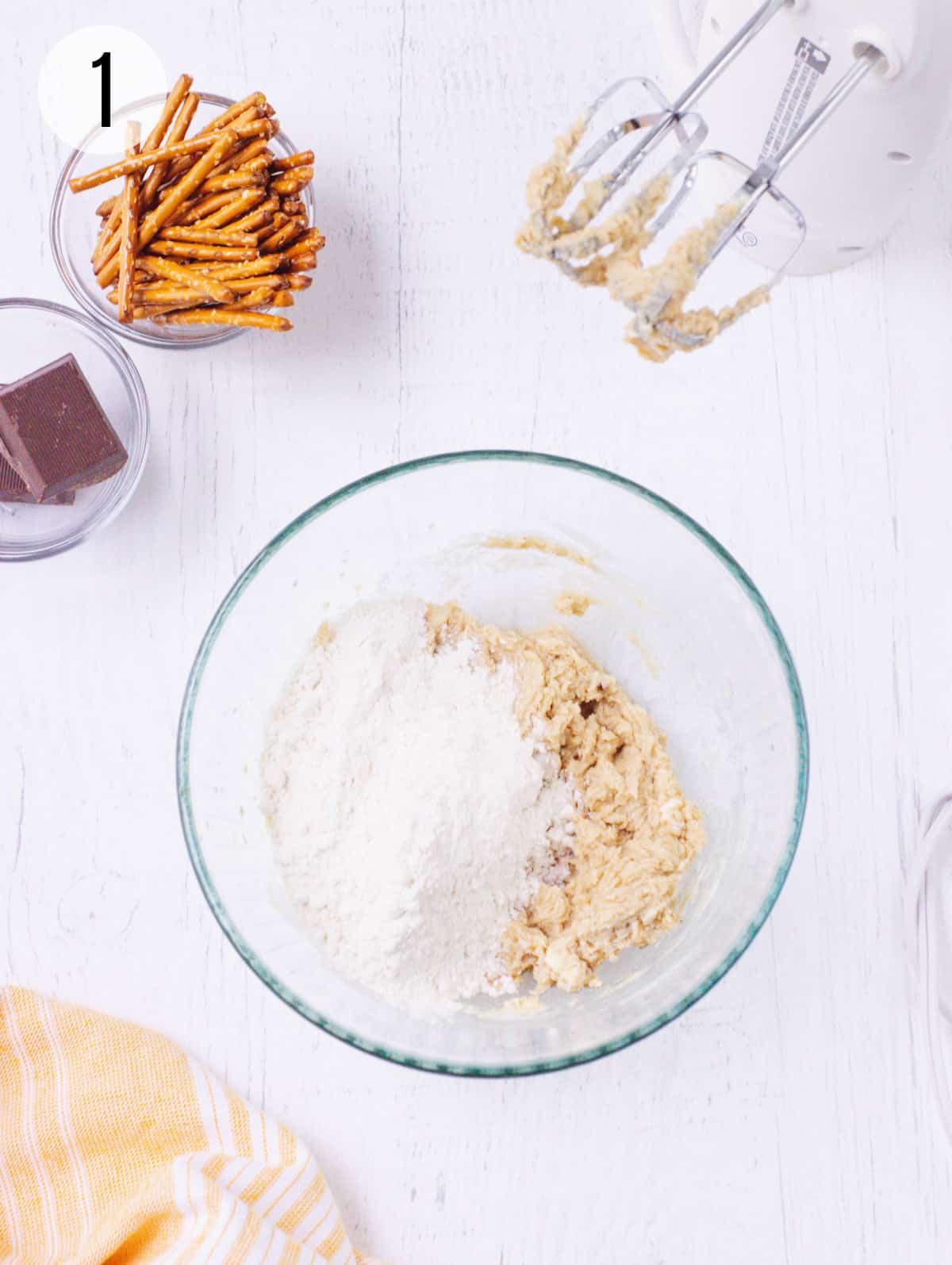 Glass bowl with creamed butter and sugar and flour mixture with chocolate squares and pretzels in upper left and electric mixer beaters in upper right.