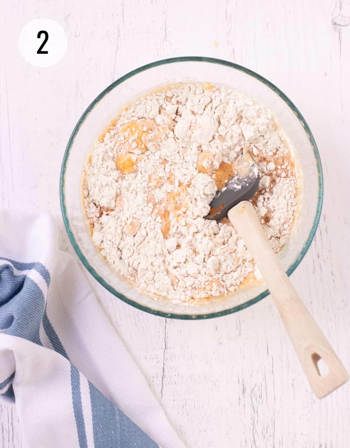 Glass bowl of flour and pumpkin mixture to make pumpkin waffles being stirred by grey rubber spatula with wooden handle and blue and white striped towel in lower corner. 