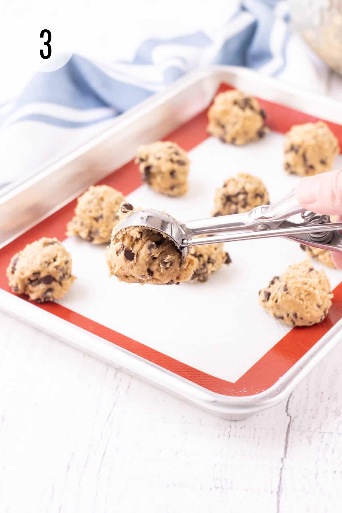 Cookie dough balls on a baking sheet lined with red and white silicone baking mat with silver scoop for dough with blue and white towel in background. 