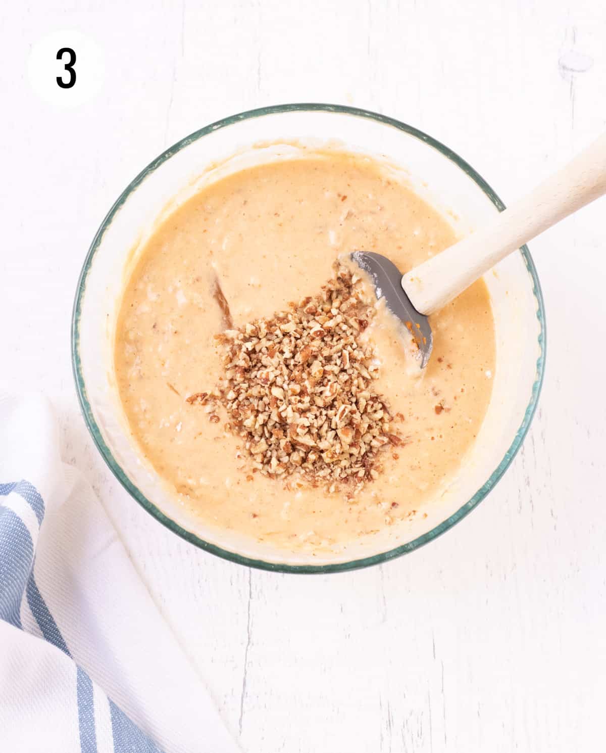 Pumpkin waffle batter in glass bowl with chopped nuts in center being stirred by grey rubber spatula with wooden spoon and blue and white striped towel in lower corner. 