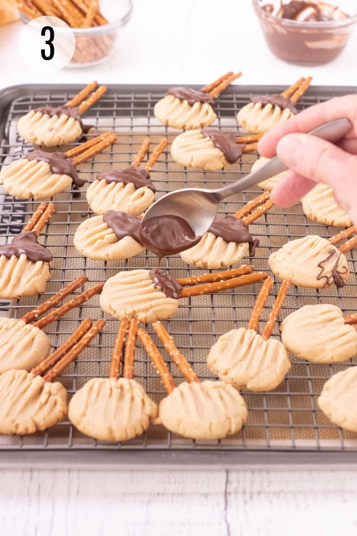 Drizzling melted chocolate on to brown sugar shortbread cookies shaped like witches brooms with pretzels sticks for broom all on a baking rack and bowls of pretzels and melted chocolate in upper background. 