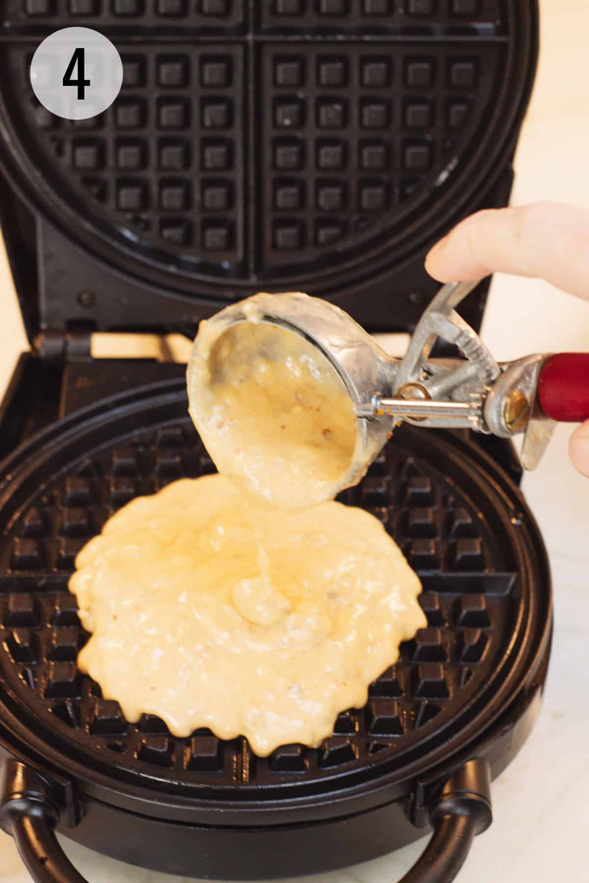 Round black waffle maker with large scoop adding pumpkin waffle batter to waffle maker. 