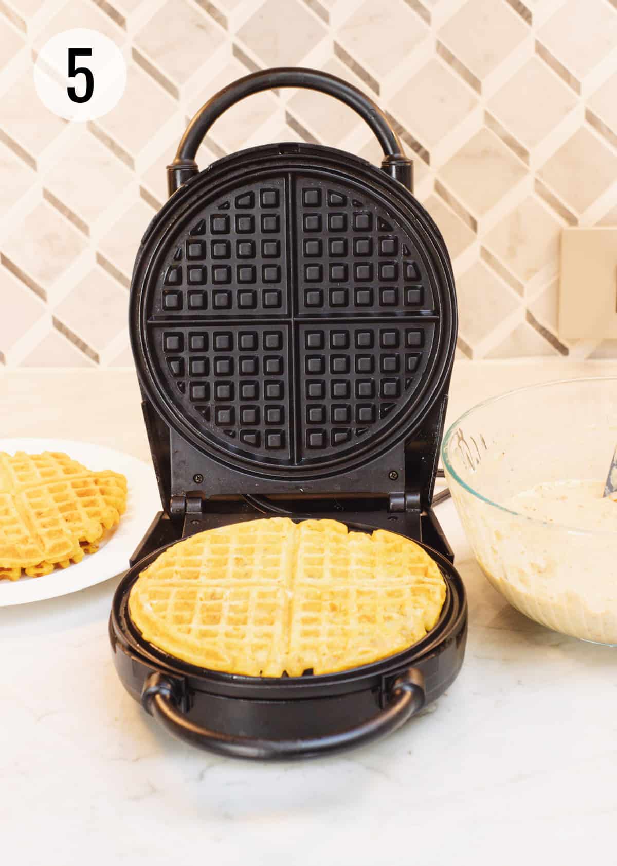 Round black waffle maker with baked pumpkin waffle on it and batter bowl to right and more baked waffles on a plate on left. 