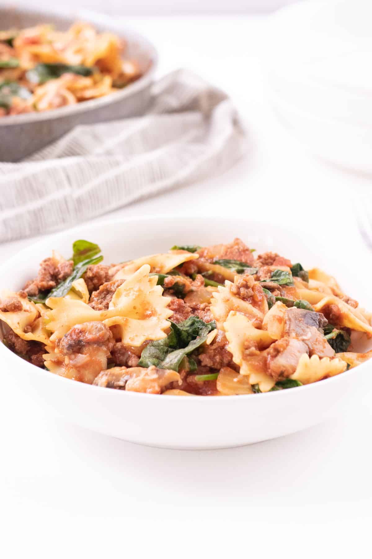 White bowl filled with stove top lasagna including ground beef, bow tie pasta, spinach and marinara sauce with skillet in upper left corner. 