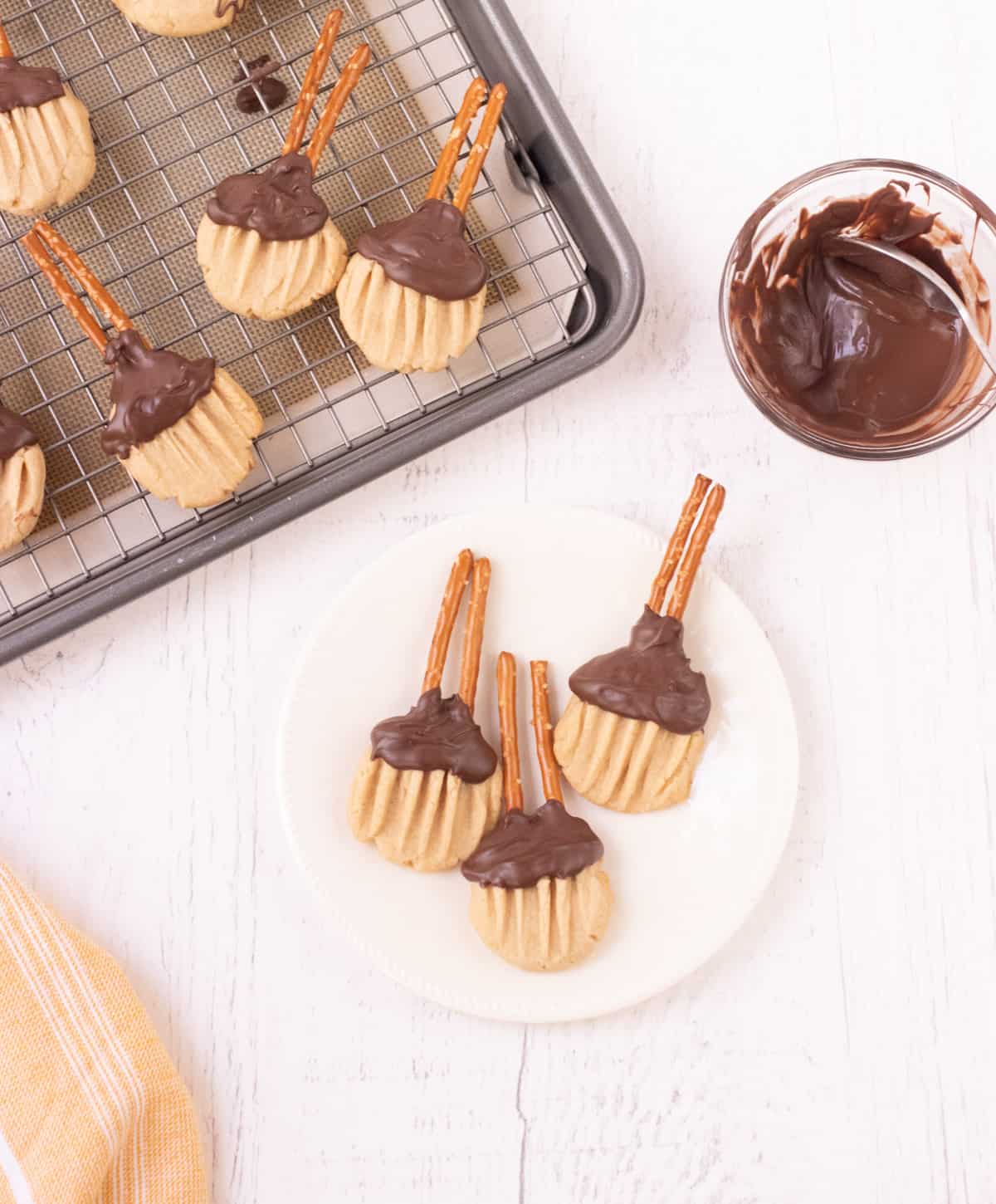 Three brown sugar cookies shaped like a witches broom with chocolate and pretzel sticks on a plate and extras on a baking rack in upper left and melted chocolate in bowl to upper right. 