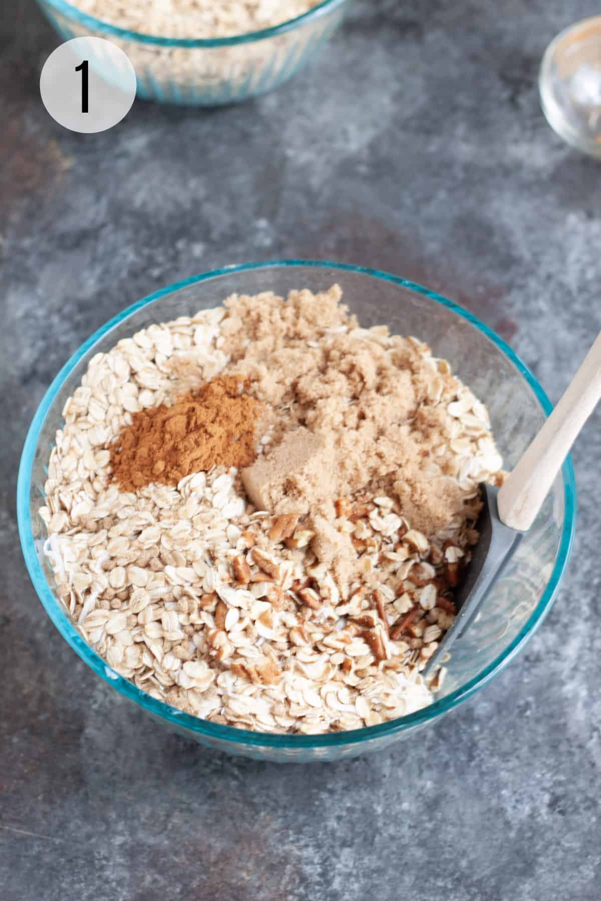 Glass bowl with ingredients for homemade granola like nuts, cinnamon, brown sugar and oatmeal with a grey rubber spatula. 