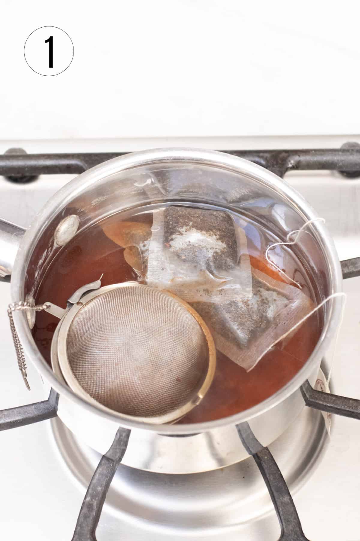 Silver saucepan with tea bags and spice ball steeping over hot stove. 