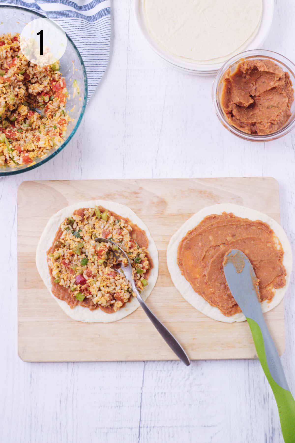 Wooden cutting board with flour tortillas spread using spoon and green and grey rubber spatula with vegetarian burrito filling and refried beans with bowls of each in upper background. 