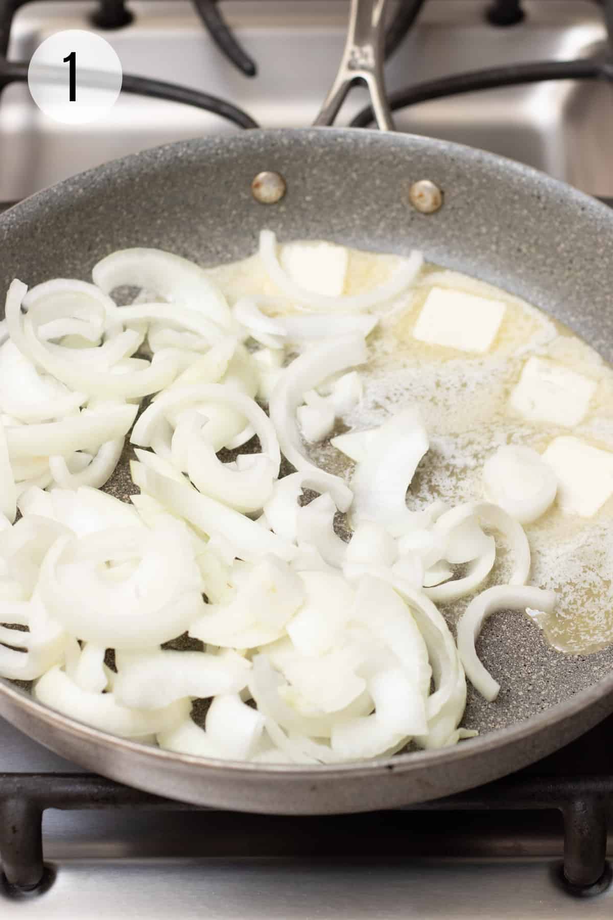 Grey skillet with sliced onions and square pats of butter sautéing on stove. 