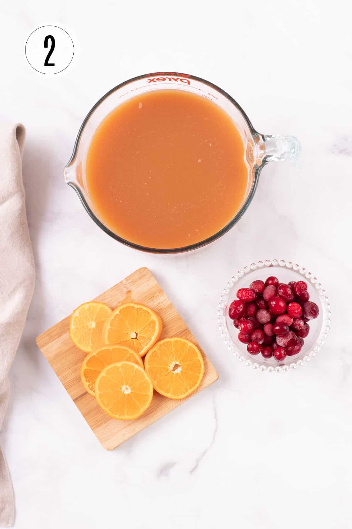Large measuring cup with orange cranberry tea concentrate with wooden cutting board of orange slices and bowl of cranberries below and tan linen on side. 