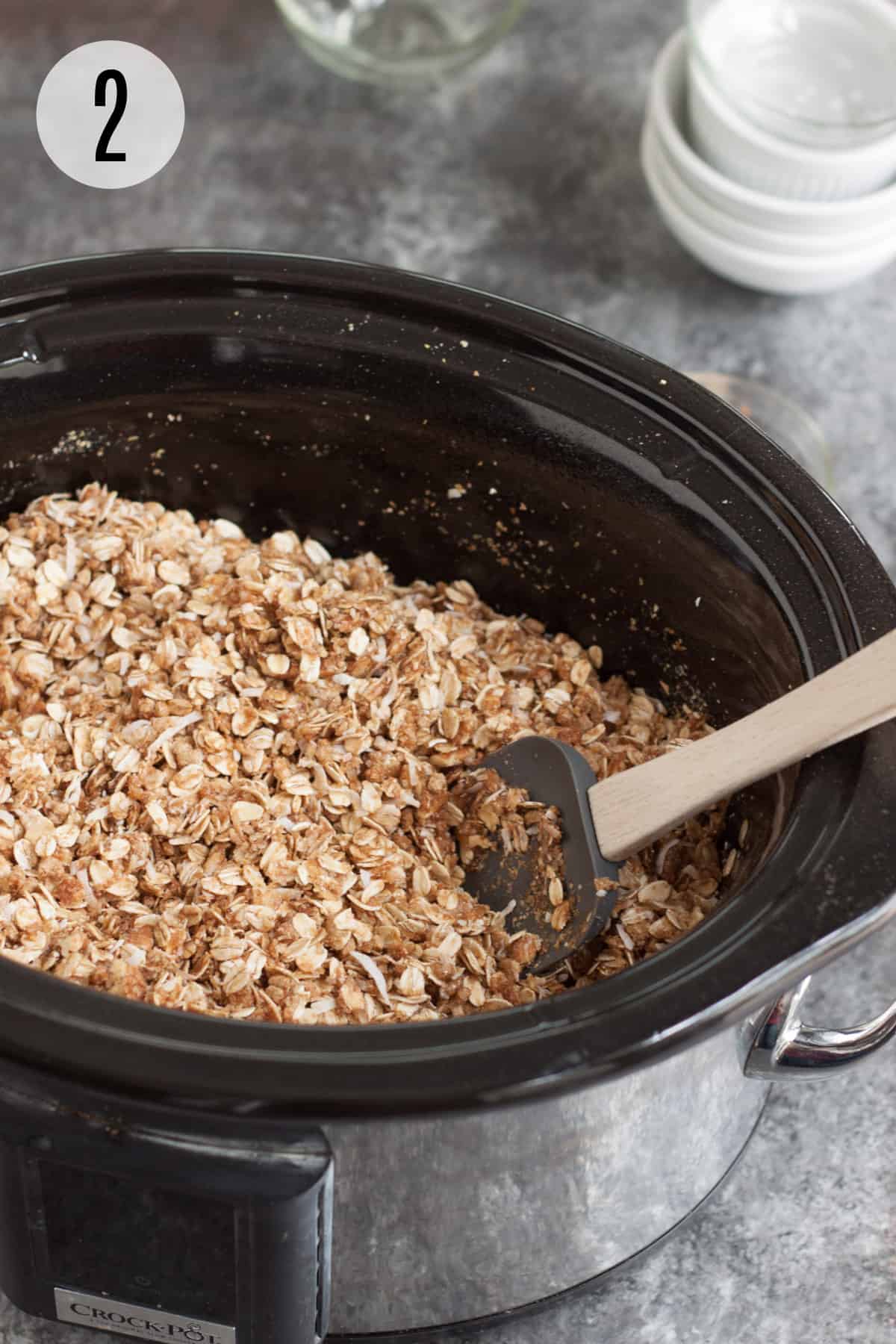 Black slow cooker with homemade granola mixture being stirred by grey rubber spatula with wooden handle and ingredient bowls in background. 