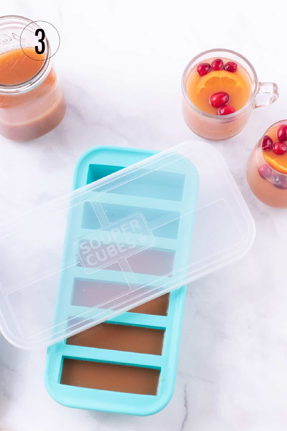 Aqua SouperCubes tray filled with Orange Cranberry Tea concentrate and jar of concentrate in upper left and glass cups of tea with cranberry and orange slice garnish in upper right. 