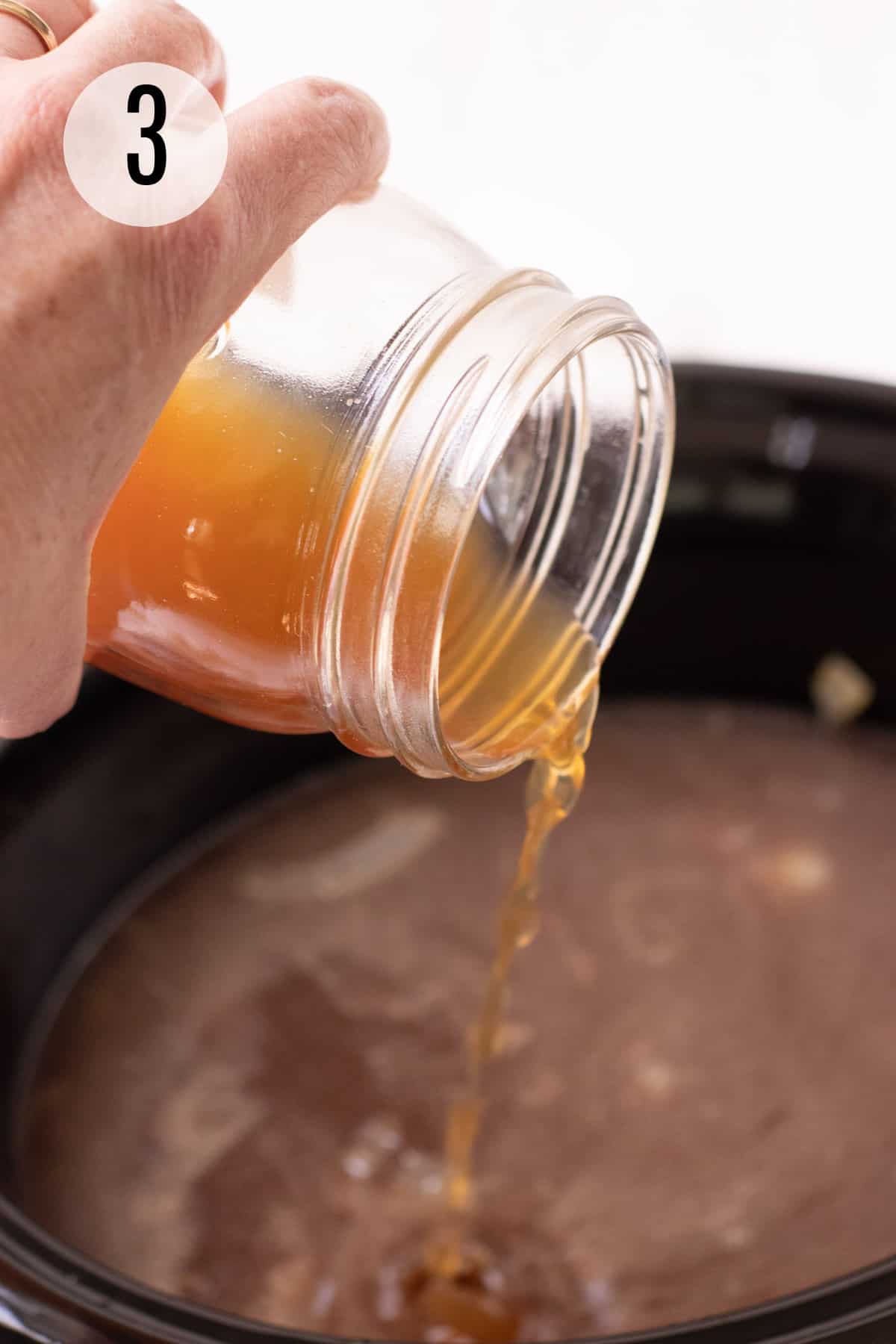 Glass jar of beef broth being poured into a black crock pot full of French Onion soup ingredients. 