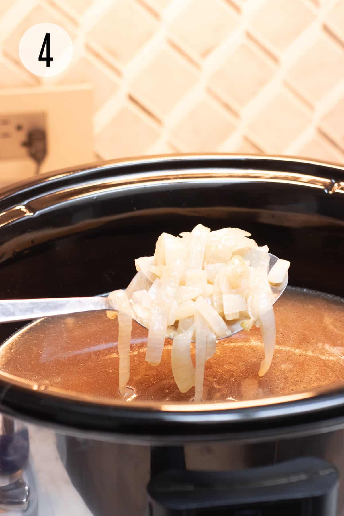 Black and silver Crock pot filled with French onion soup with silver spoon scooping out sliced onions. 