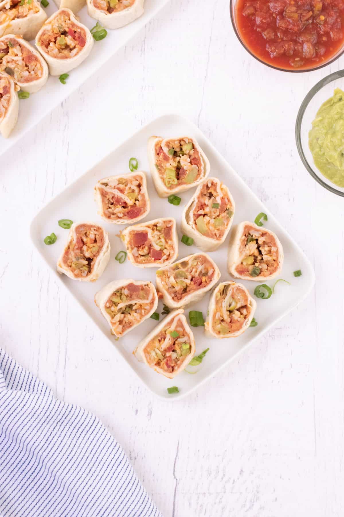 White plate with vegetarian burrito bites garnished with sliced green onions with platter of more burrito bites and bowls of guacamole and salsa in upper background and blue striped towel in lower left corner. 