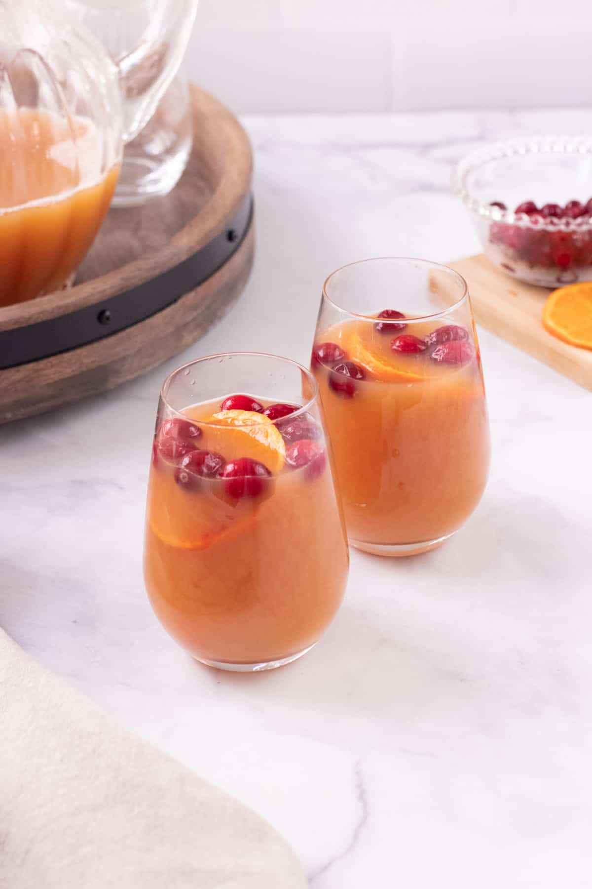 Two glasses of Orange Cranberry Tea with cranberries and orange slices floating as garnish and pitcher of tea mixture and bowl of cranberries and orange slices in background. 