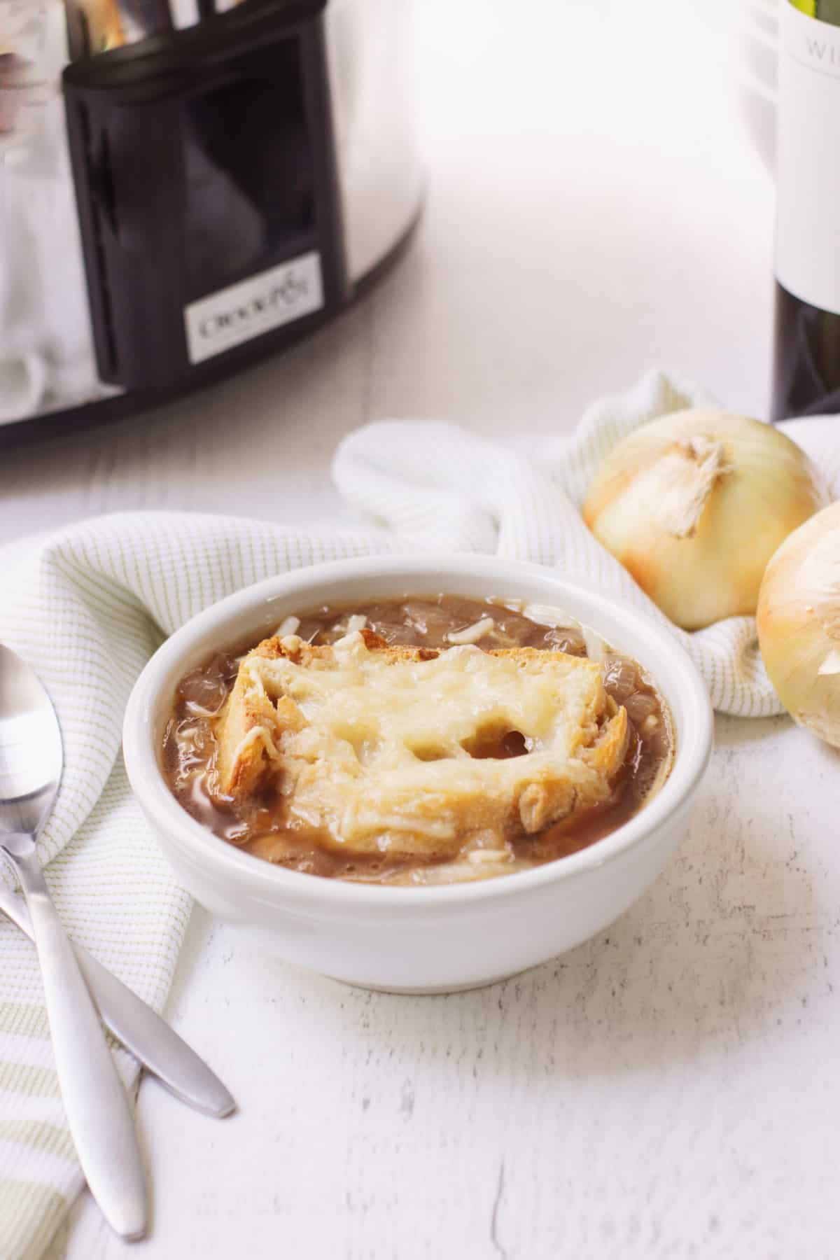 white bowl with French onion soup topped with crust bread and cheese with crockpot in upper left, spoons to left and onions to right of bowl. 