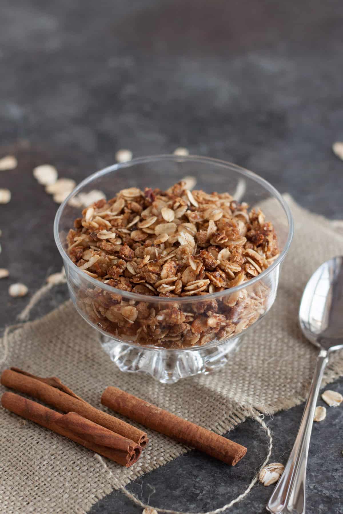 Glass bowl with homemade granola on top of a burlap napkin with cinnamon sticks and silver spoon.