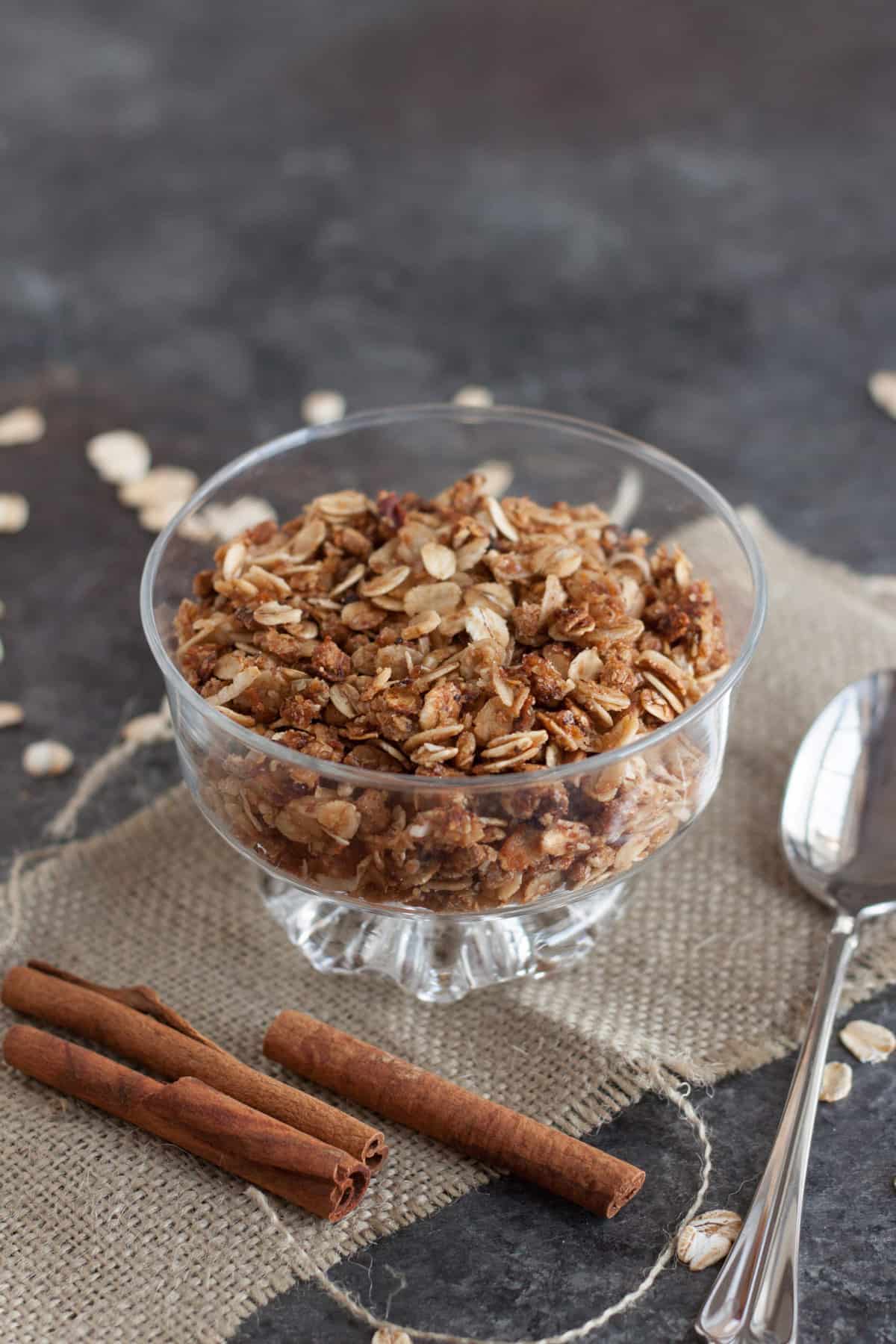 Glass bowl with homemade granola on top of a burlap napkin with cinnamon sticks and silver spoon. 