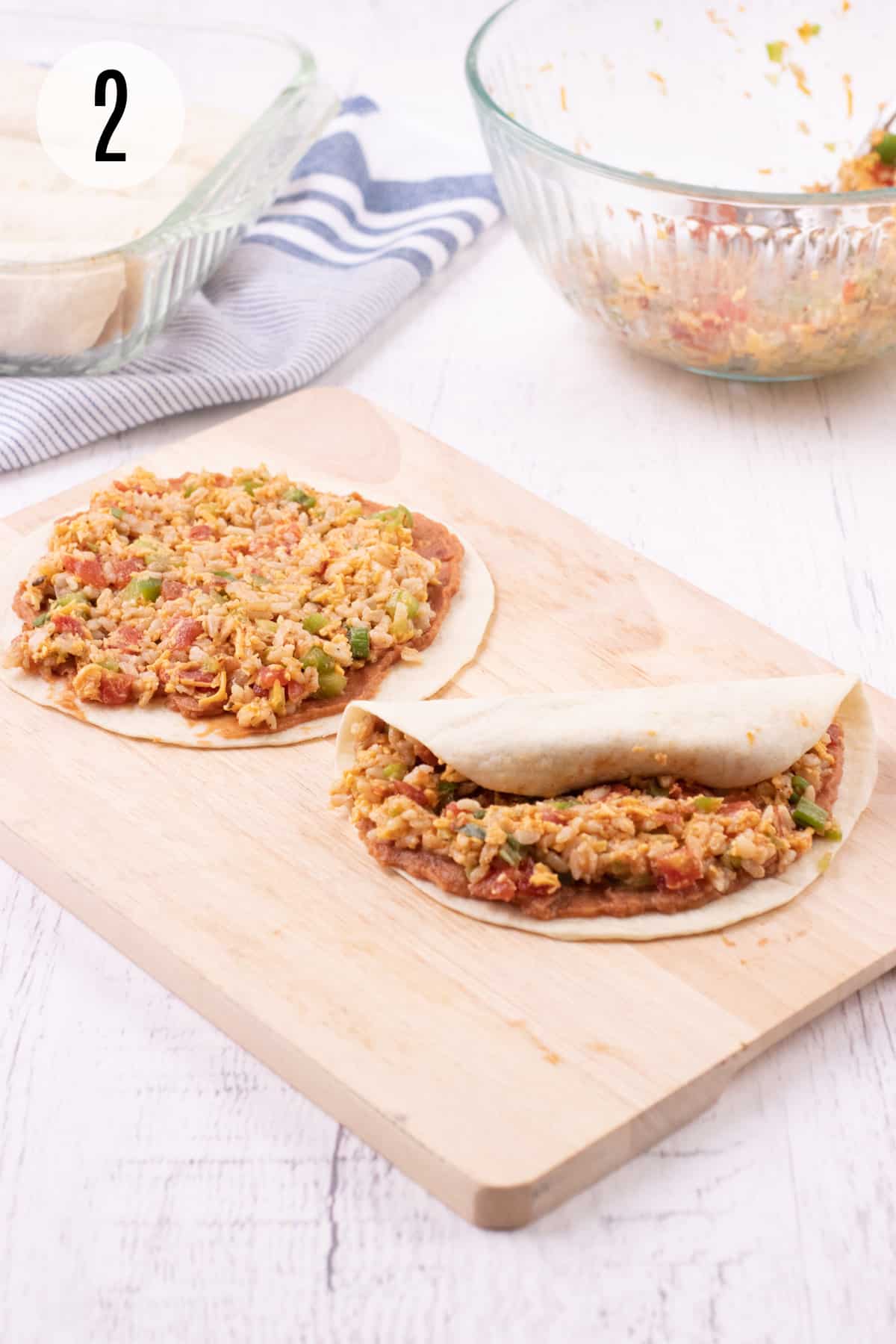 Wooden cutting board with tortillas filled with burrito bite filling and partially rolled up with pan of rolled tortillas and glass bowl of mixture in upper background. 