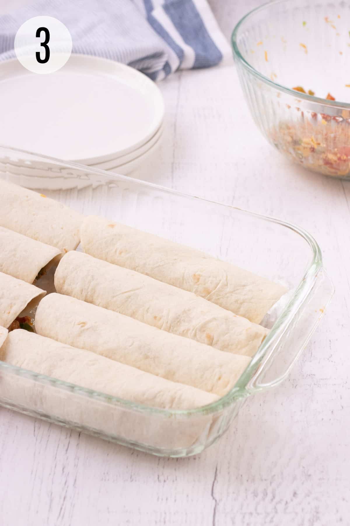 Glass baking dish with rolled tortillas filled with burrito bite mixture and white plates, blue and white striped towel and glass bowl of mixture in upper background. 