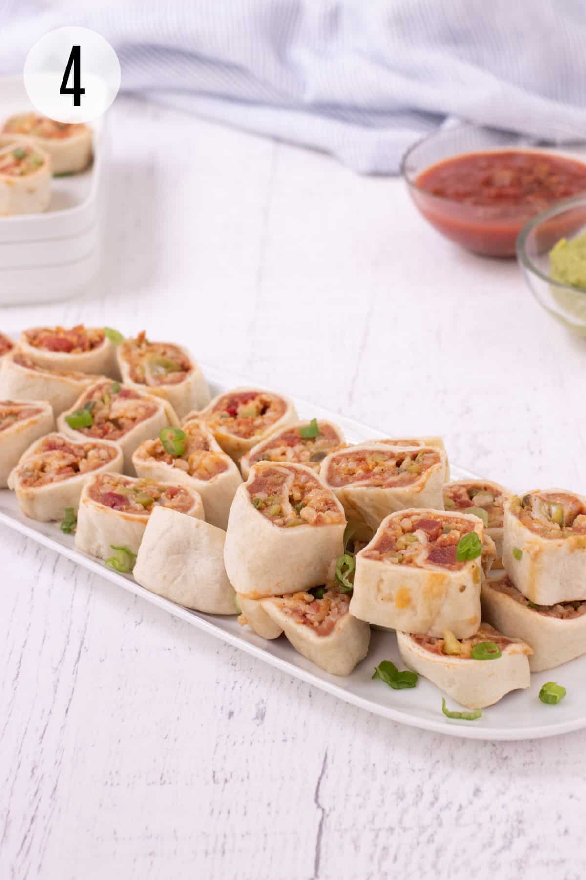 Platter with stacked burrito bites garnished with sliced green onions and plate of more bites and bowls of guacamole and salsa in upper background. 