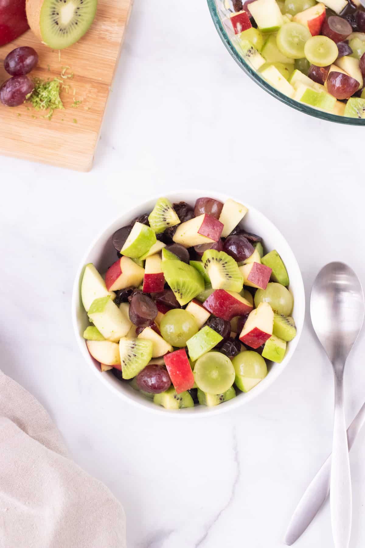 Top view image of holiday fruit salad in white bowl with red and green apples, red and green grapes, kiwi and dried cherries with wooden cutting board and glass bowl in upper right and left corners. 