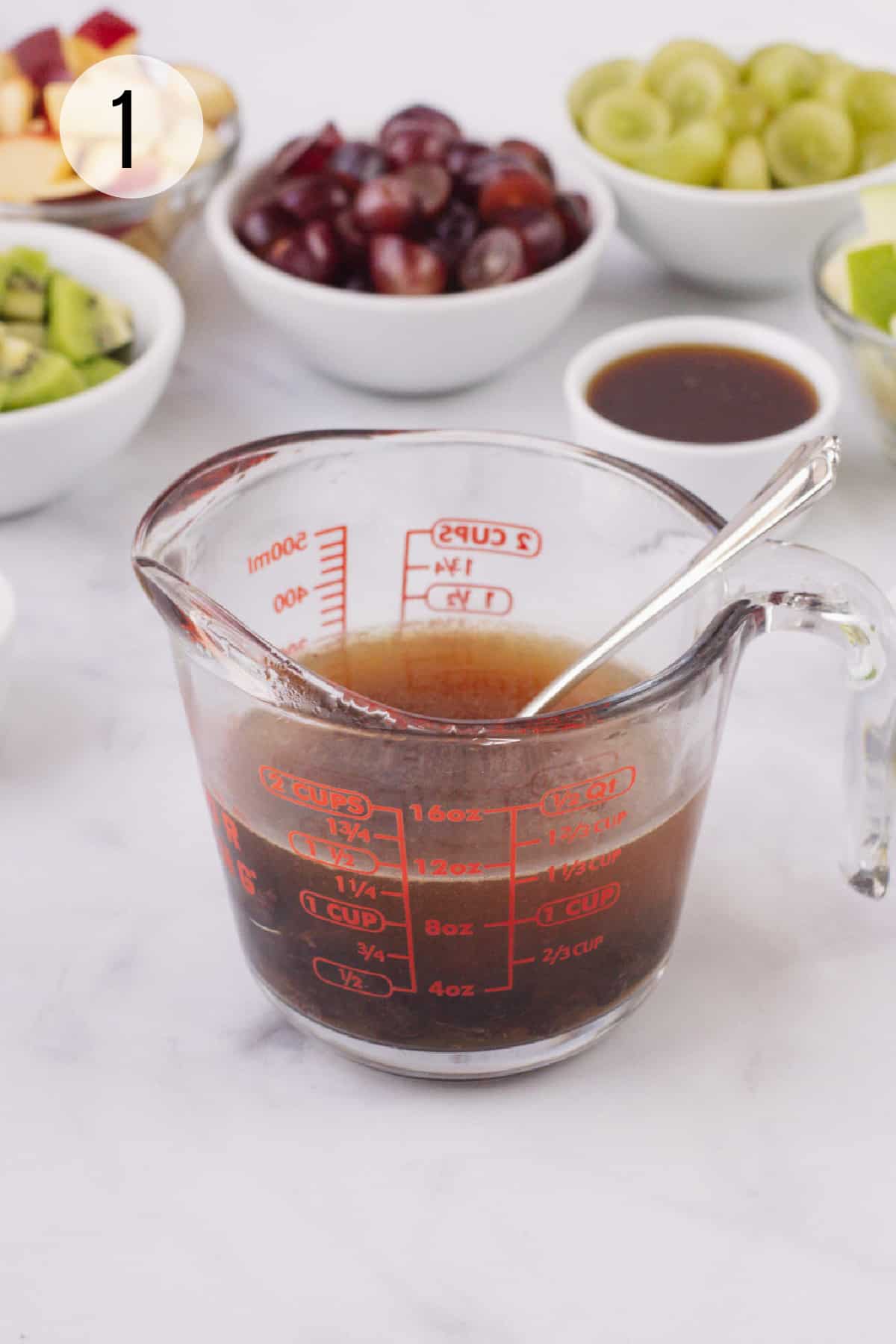 Glass measuring cup with water and dried cherries soaking and ingredients for Holiday Fruit Salad in bowls in background.