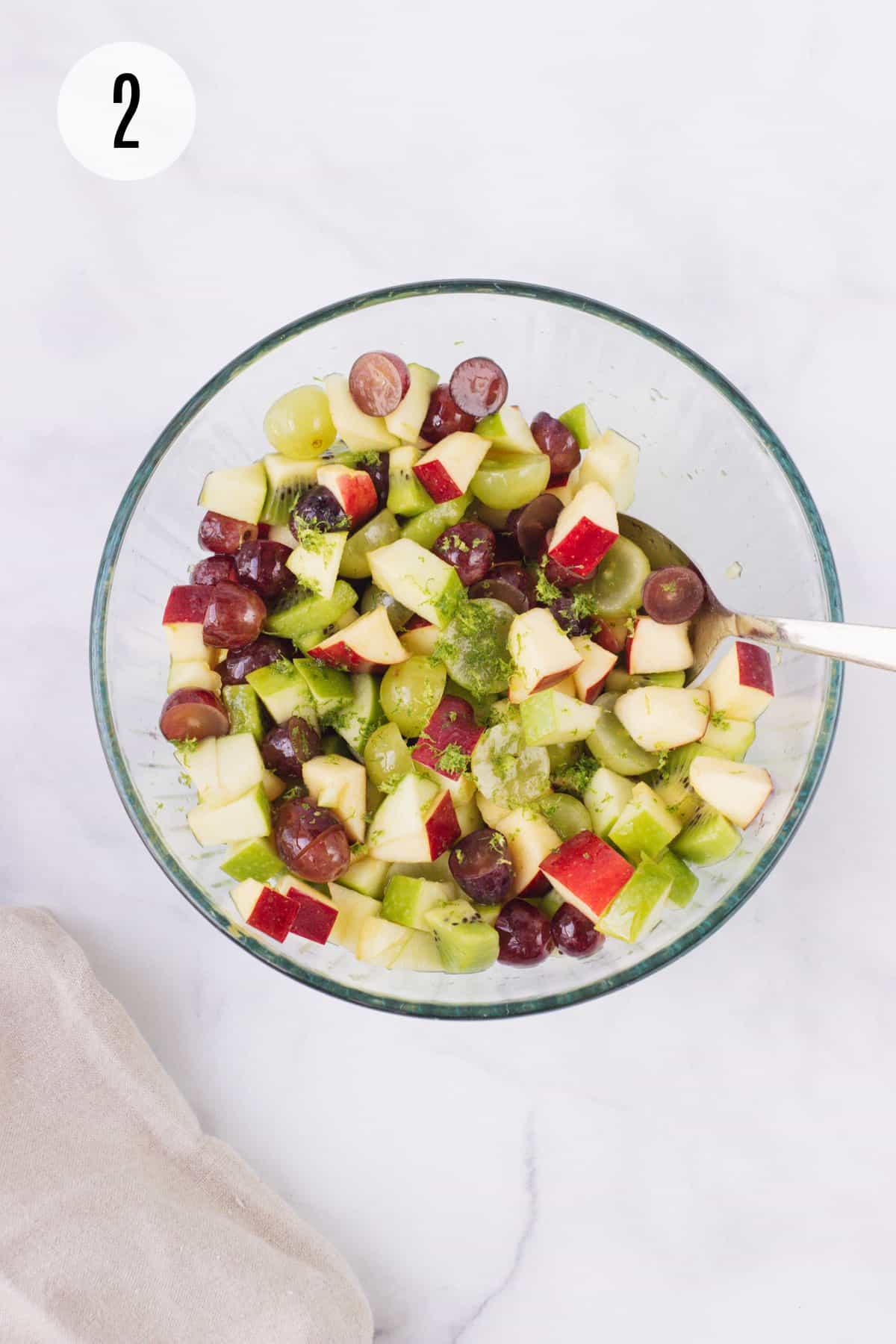 Top view image of Holiday Fruit Salad with chopped apples, grapes, kiwi and lime zest in a glass bowl being stirred by silver spoon. 