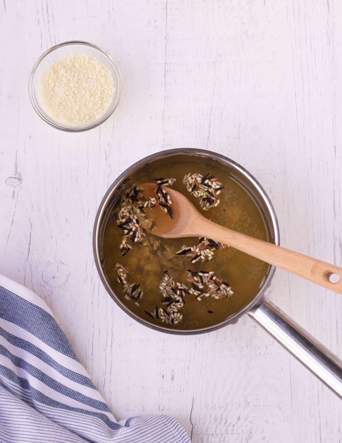 Top view of chicken broth and wild rice in a metal saucepan with a wooden spoon stirring and bowl of grated parmesan cheese in upper left and blue and white towel in lower left.