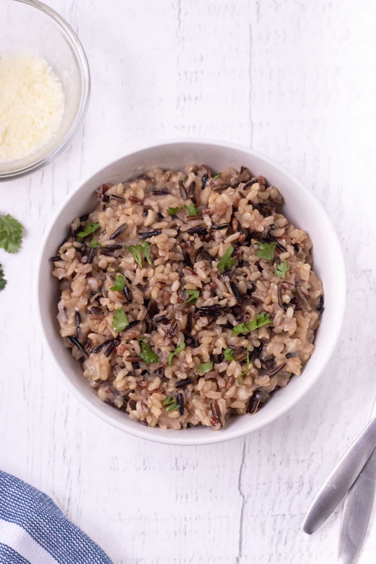 Top view of brown and wild rice in a white bowl with parsley for garnish and a bowl of grated parmesan cheese in upper left corner.